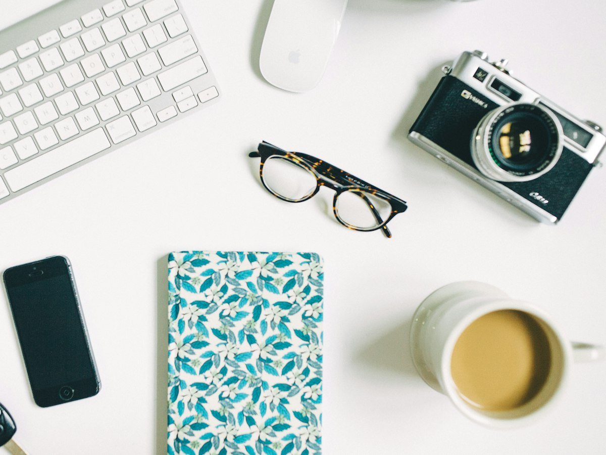 A keyboard, a camera, glasses, a notebook and a cup of coffee laid out on a white desk